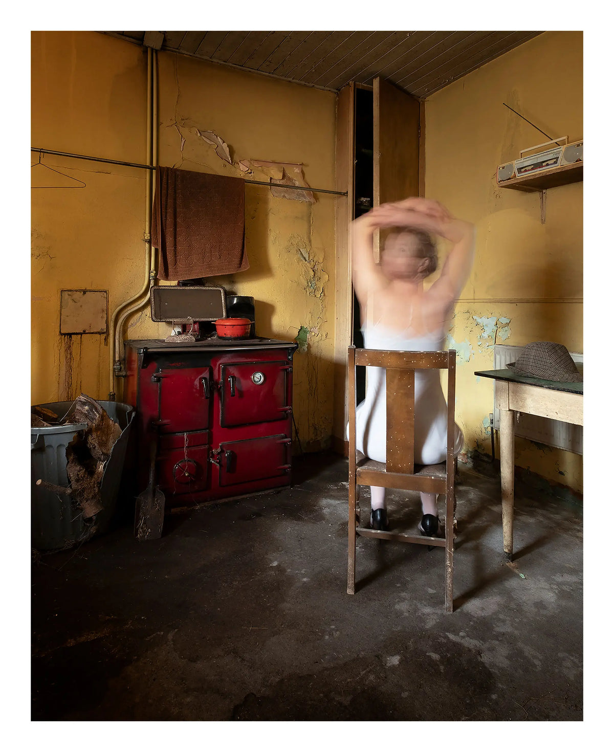 Woman in a white nightdress with arms raised in motion blur beside a red vintage stove in an aged yellow kitchen.