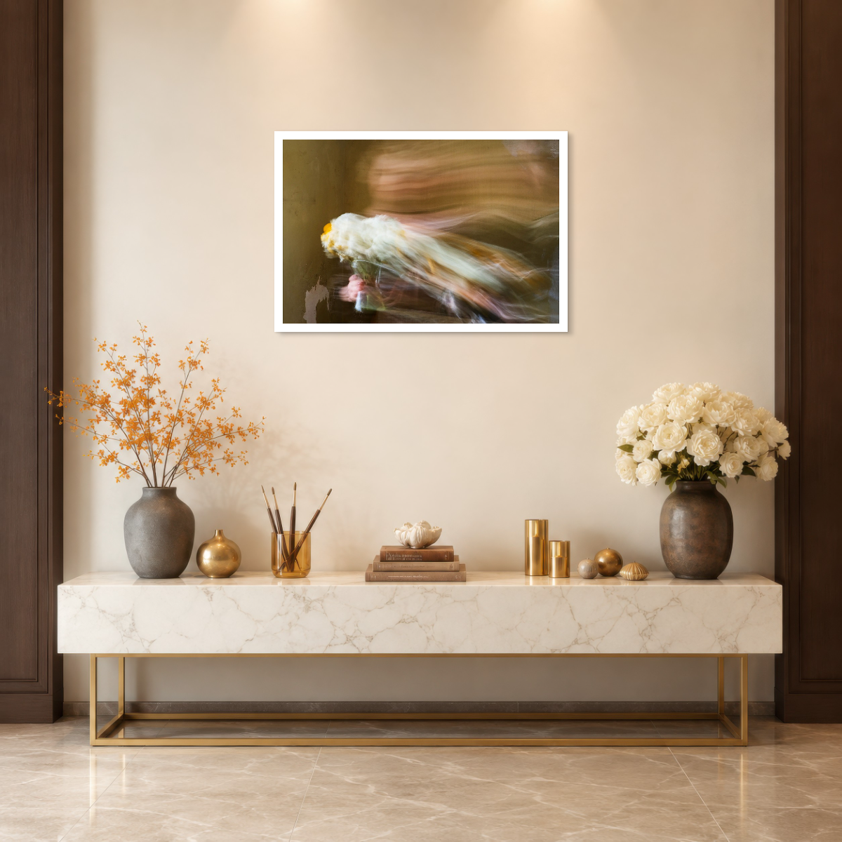 A framed abstract art print hangs on a beige wall above a marble console table decorated with vases, gold accents, and books. The print features a soft, blurred composition with flowing tones of white, gold, and dark brown.