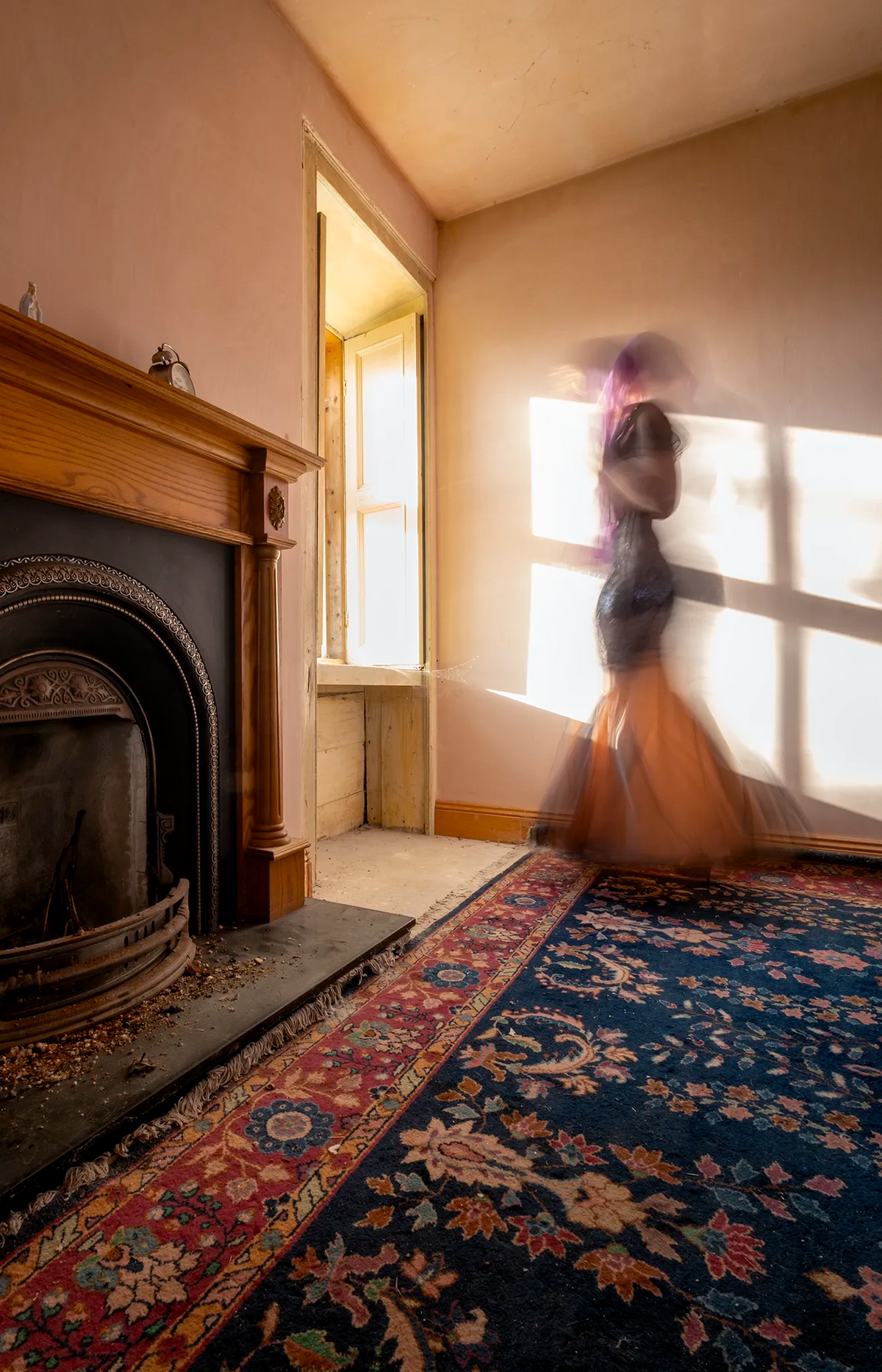 Blurred female figure in a dark dress standing at a window in a faded pink room of an old Georgian house in Ireland, illuminated by warm afternoon sunlight.