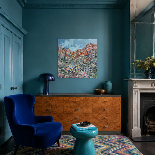 A colorful interior room scene featuring a landscape painting hanging on a teal wall above a wooden cabinet, with a blue velvet armchair, turquoise side table, and fireplace visible.