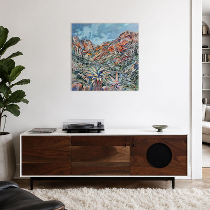 A square contemporary painting on canvas depicting a colorful desert landscape with mountains and palm trees, displayed on a white wall above a modern wooden media console in a living room setting.