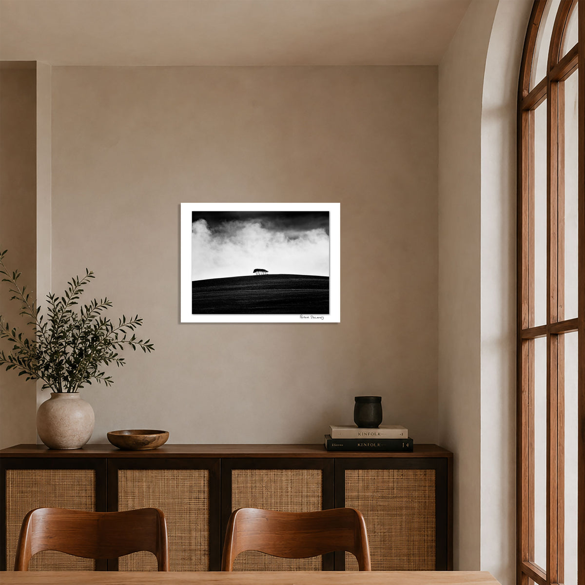 Peter Delaney’s black-and-white landscape photograph of a lone group of trees on a dark hillside, framed in white and displayed above a wooden sideboard in a neutral contemporary interior.