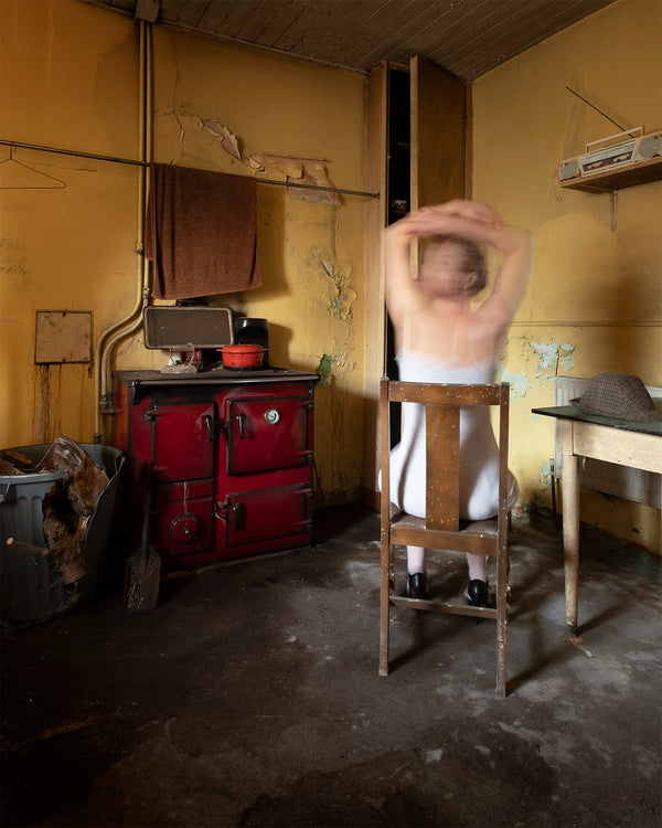 Seated Woman in the Kitchen by Robyn Daly, a long-exposure self-portrait of a woman in a white nightdress sitting on a wooden chair in a worn yellow kitchen.