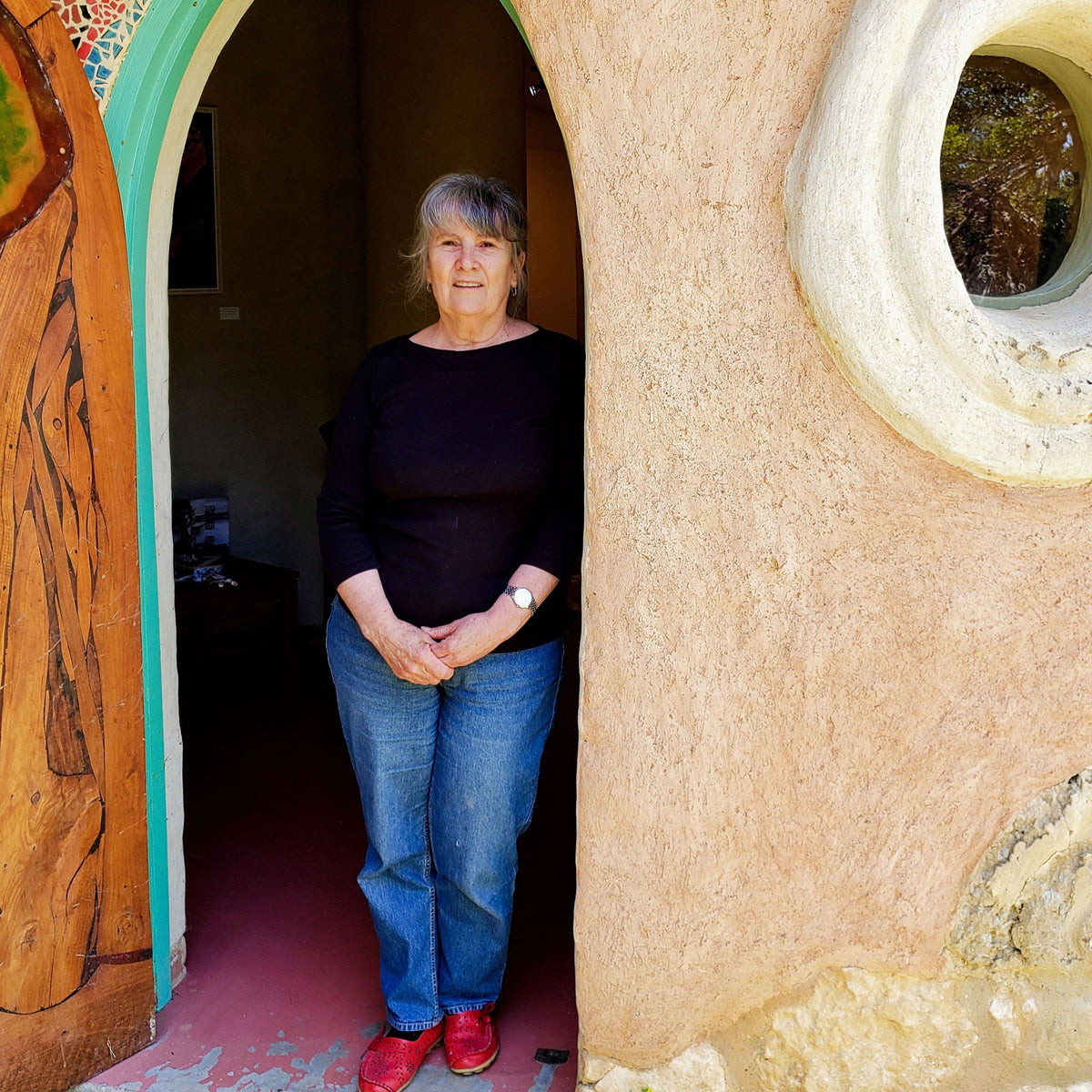 Australian painter and artists Karen Hammat stands in a doorway wearing a black long-sleeve top, blue jeans, and red sneakers, posed in front of a textured beige stucco wall with a circular window.