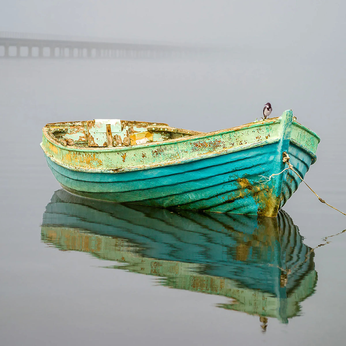 A weathered turquoise wooden rowboat floating on calm water with its reflection visible below. A small bird stands on the bow, and a faint bridge is faintly visible in the misty background.