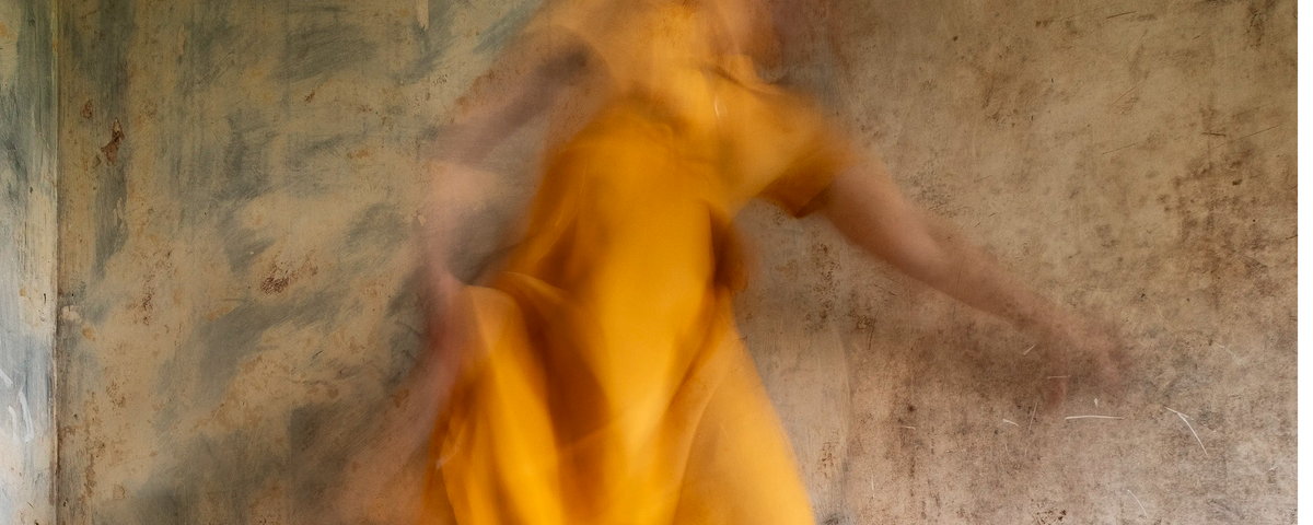 Close up of a woman in a yellow dress moving across a worn interior, her body blurred by long-exposure photography.