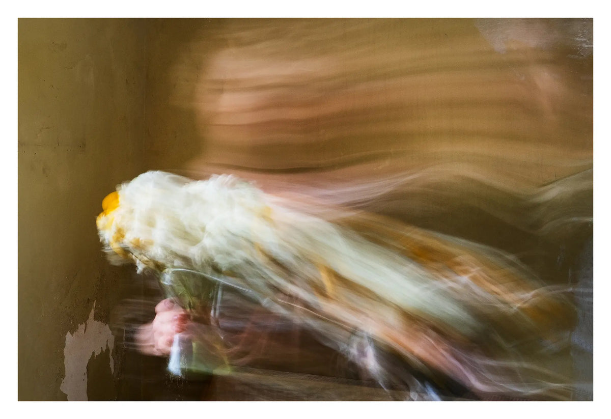 Woman holding a vase of white and yellow flowers, her figure and the flowers sweeping across the frame in long-exposure motion blur against an olive green interior wall.

