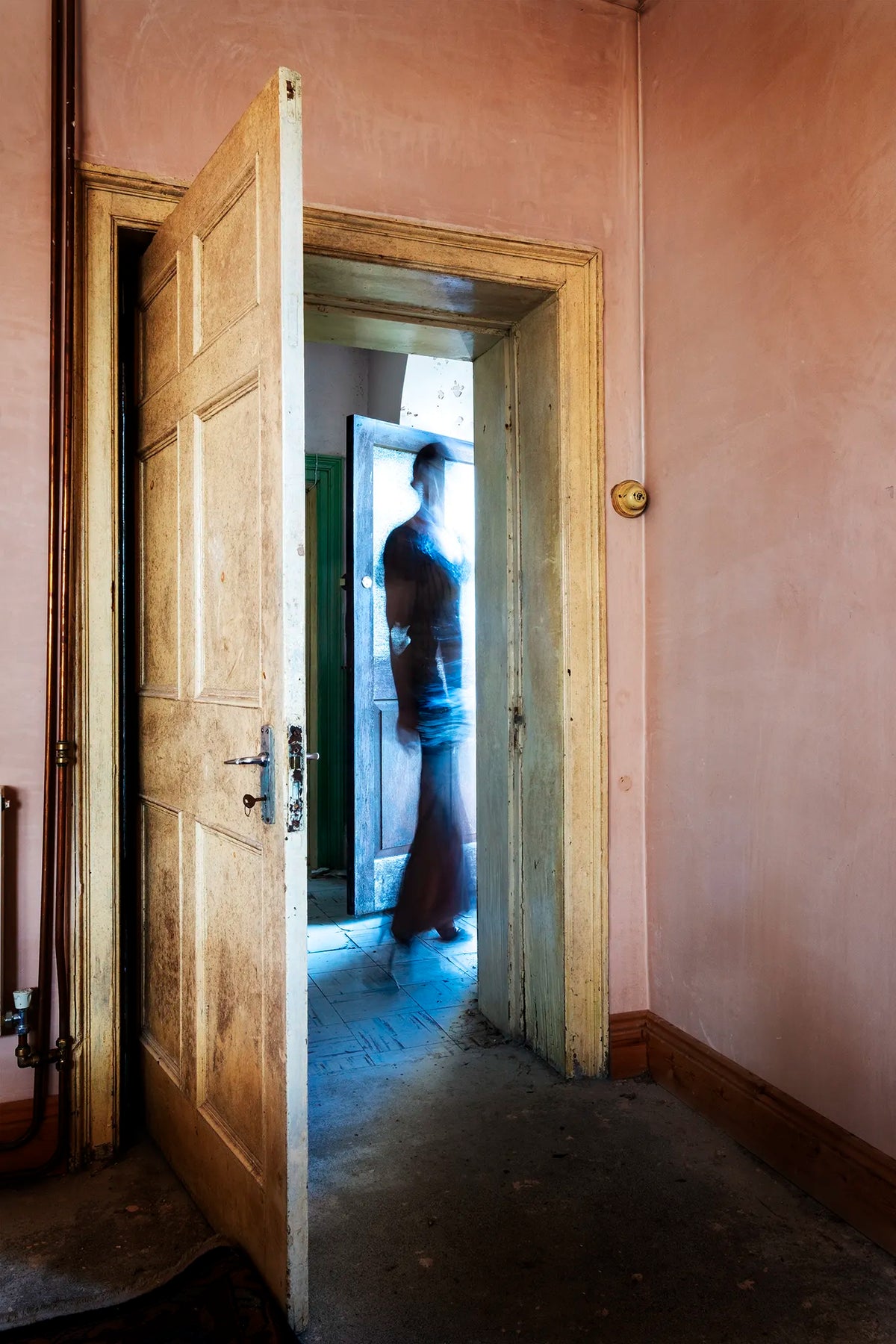 Blurred female figure in a dark dress moving through a blue-lit doorway in the faded pink interior of an old Georgian house in Ireland.